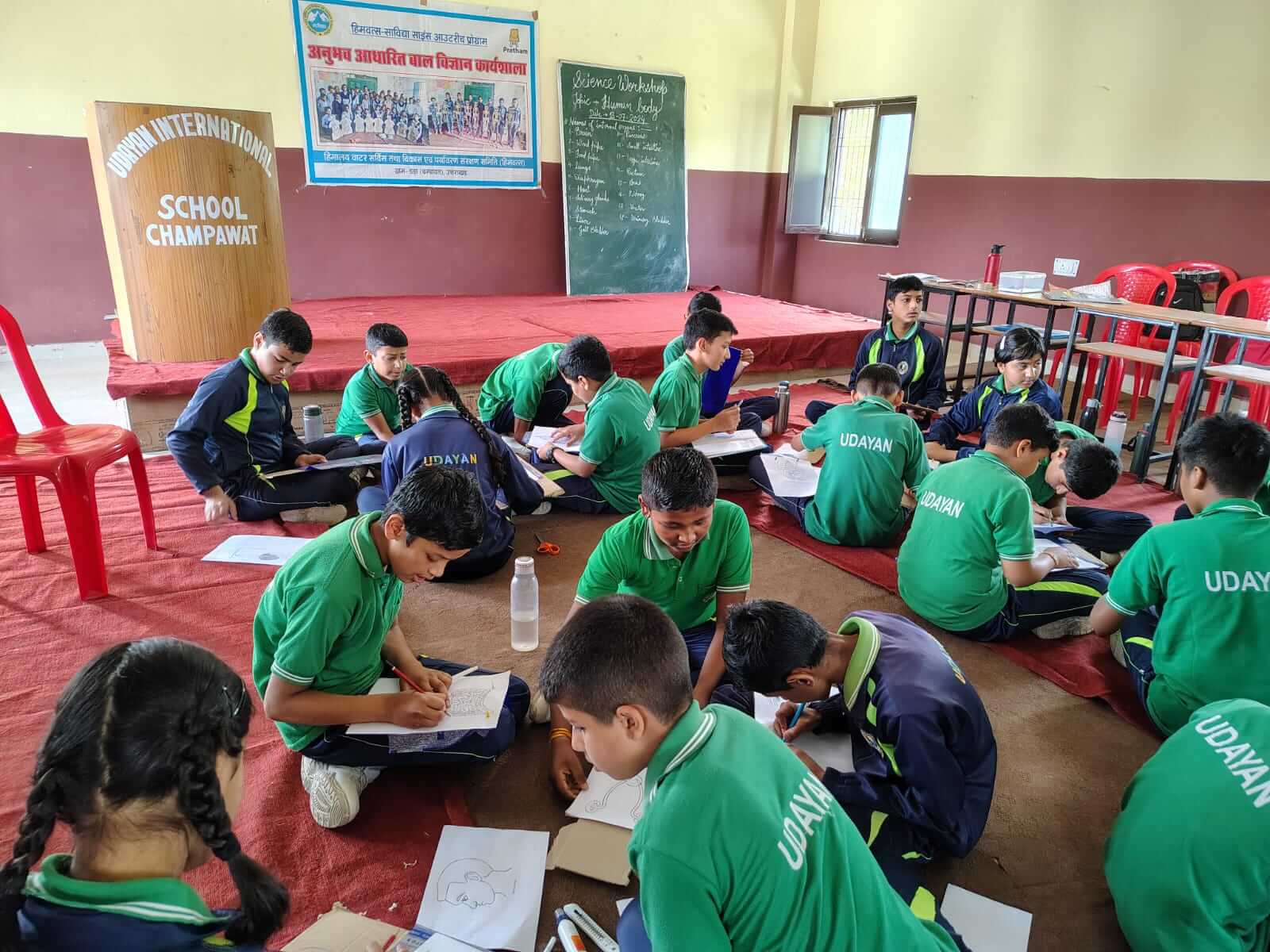 Students seated on the floor of a hall, writing, during a competition.