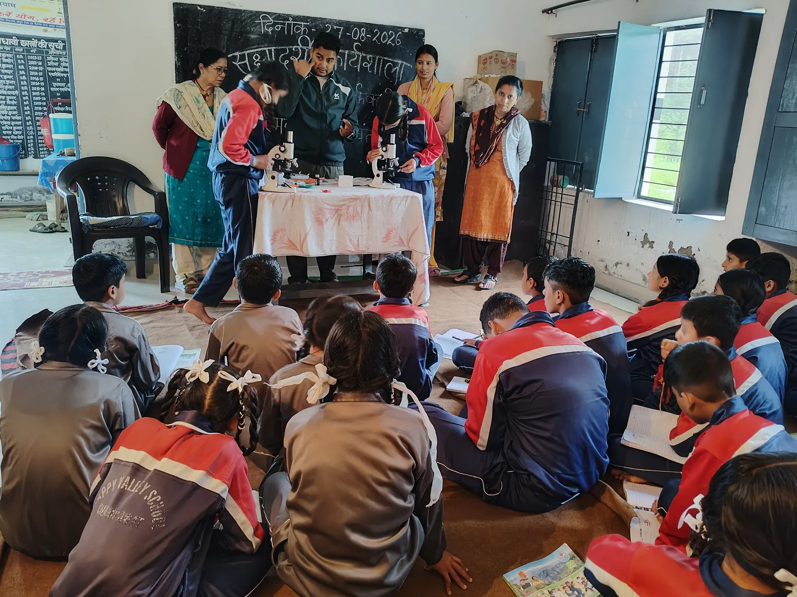 Two pupils bend over microscopes on a cloth-covered table while three teachers stand behind them, the rest of the class sitting on the floor.