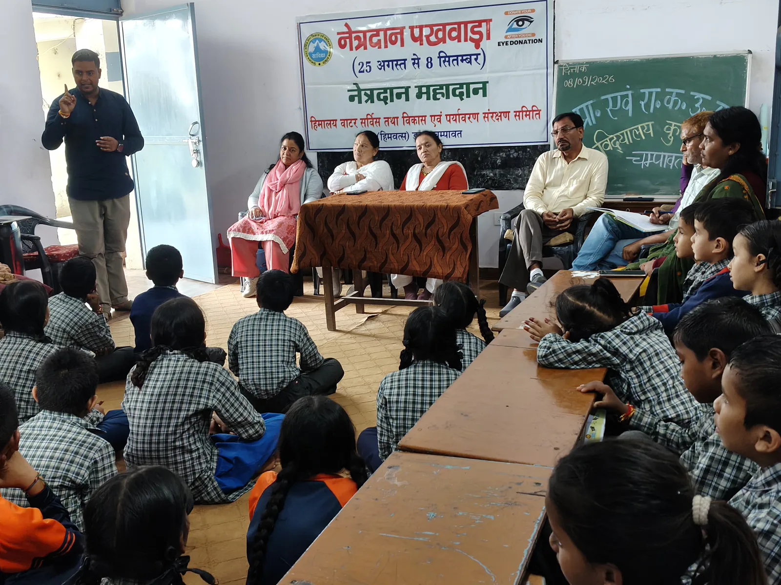 A speaker addresses pupils sitting on the floor beneath a banner reading नेत्रदान पखवाड़ा, with the date chalked on the board behind.