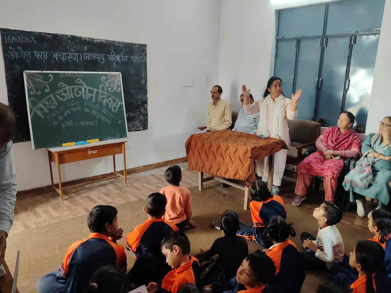 A speaker addresses children seated on the floor beside a green board headed विश्व ओजोन दिवस and dated 16/09/2026.