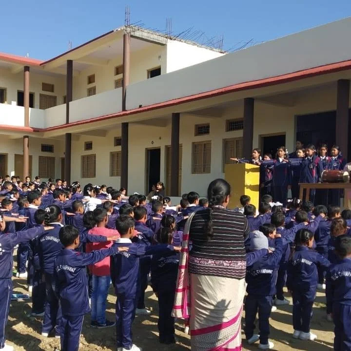 Pupils in navy tracksuits marked M.I.C. Champawat standing in rows at morning assembly, arms outstretched, in the courtyard of the two-storey school building.