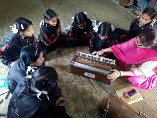 Girls gathered around a harmonium as one of them plays.