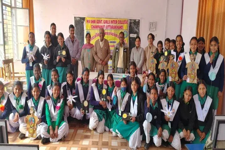 A large group of girls in green and white uniform holding models they have made, with teachers behind them under a banner reading PM Shri Govt Girls Inter College Champawat.